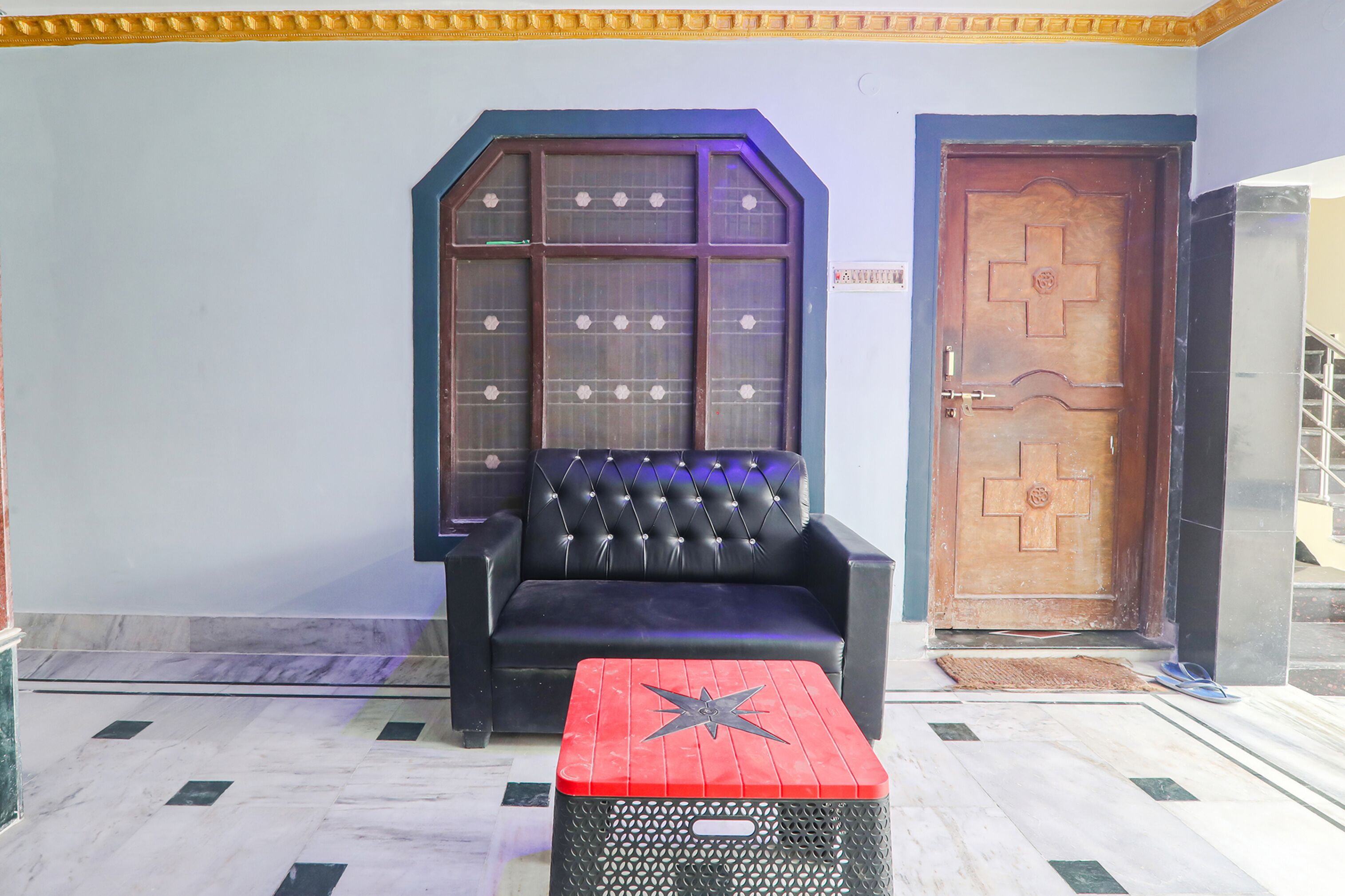 A room with a black leather sofa, a red and black ottoman, a large window with a grid pattern, and a wooden door with a cross design.
