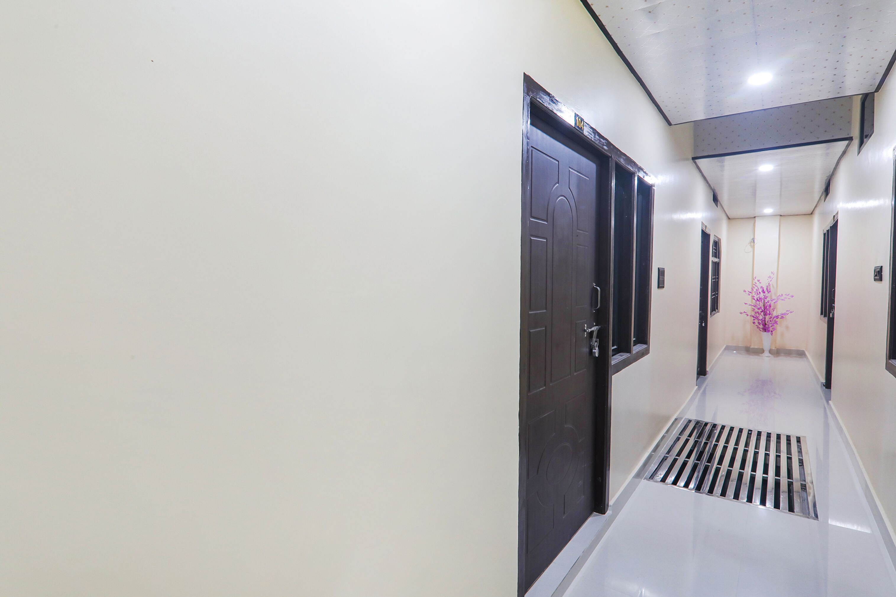 A hallway with a black door on the left, a vent on the floor, and a pink flower in a vase on the right.