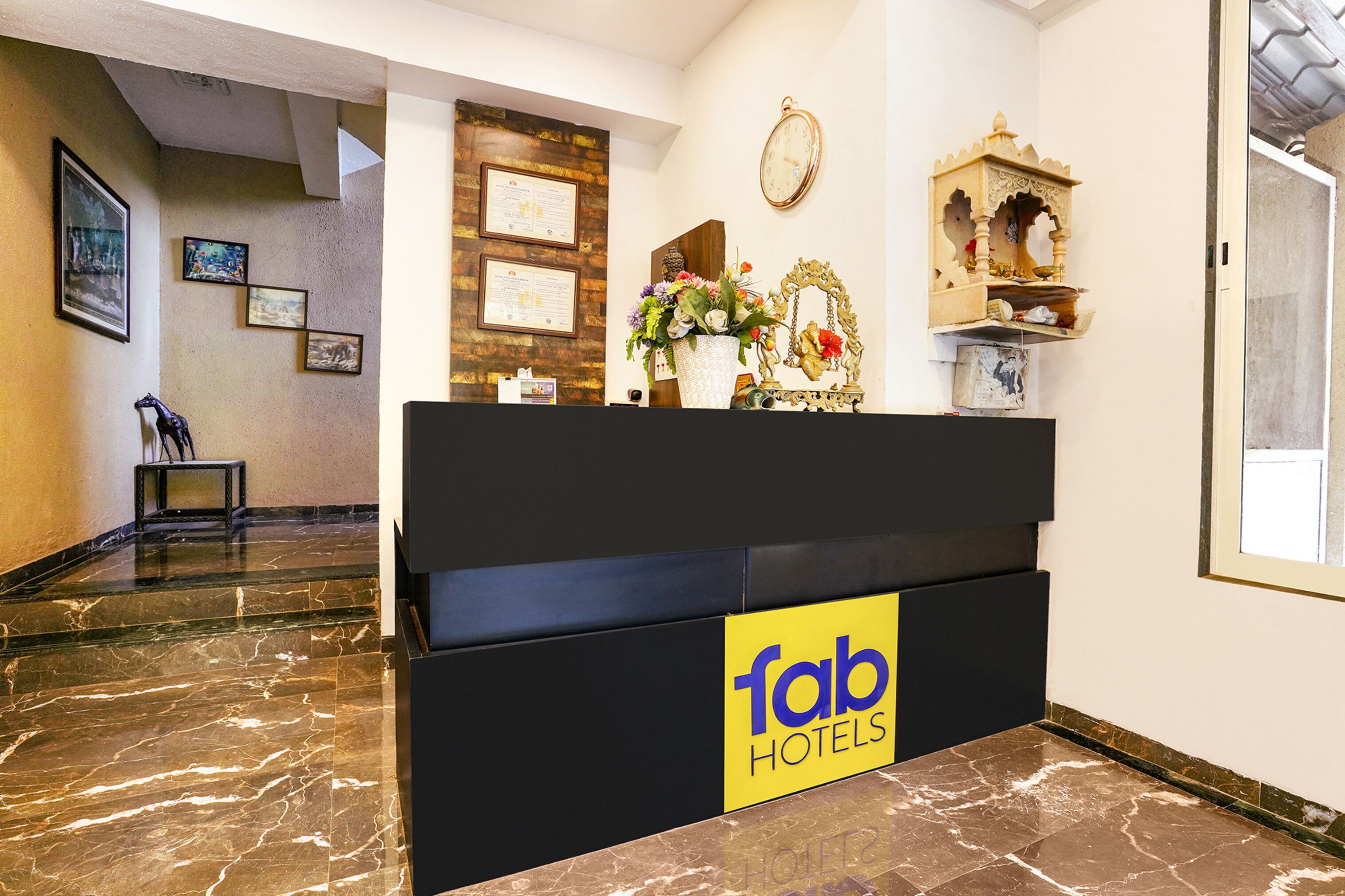 A hotel reception area with a marble floor, a reception desk with a 'Tab Hotels' sign, framed certificates on the wall, and a decorative shelf with a clock and ornaments.