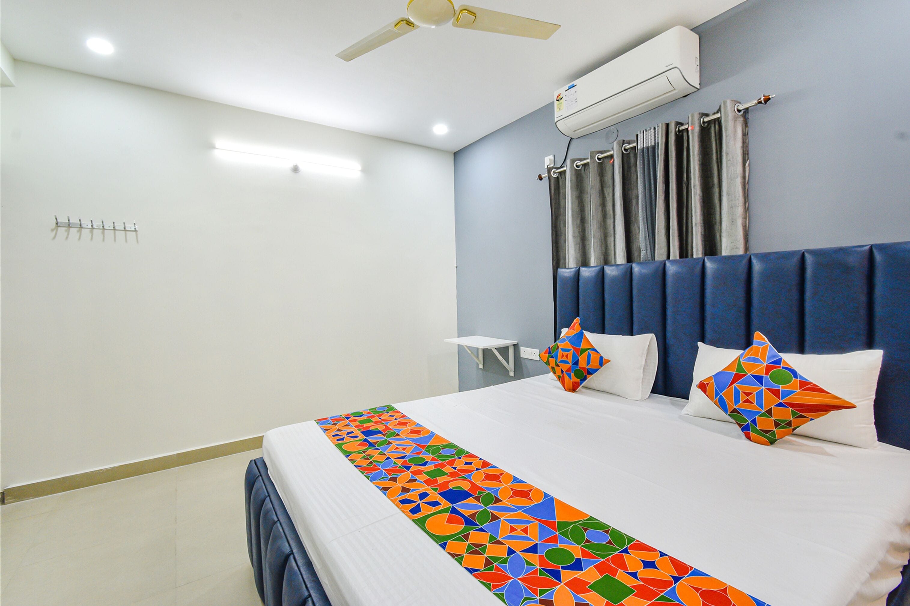 Une chambre d’hôtel avec un lit, un ventilateur de plafond, un appareil de climatisation, une petite table et une fenêtre avec des rideaux.