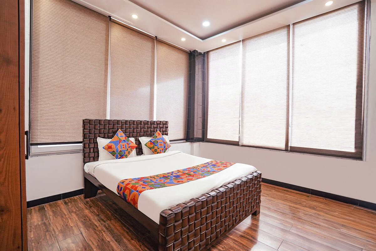 Un dormitorio moderno con cabecera de madera, una cama grande con colcha de diseño geométrico y ventanas amplias con persianas.