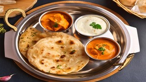 Un plato de acero inoxidable con pan naan, dos tipos de curry y un pequeño cuenco de yogur.
