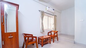 A room with a wooden cabinet, two wooden chairs, a small table, a window with curtains, and an air conditioning unit. - Fabhotel Crosscut (Coimbatore)