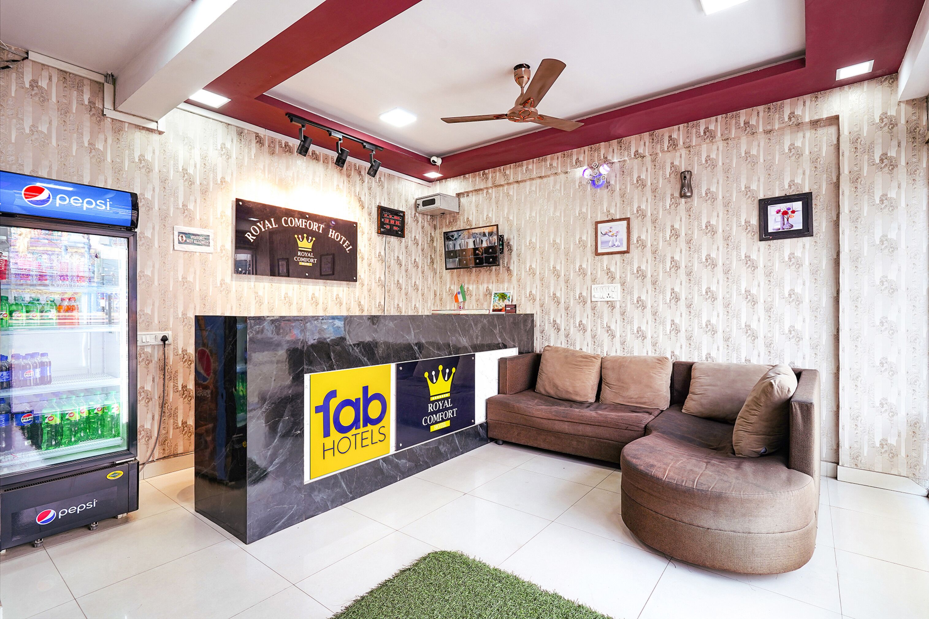 Zona de recepción de hotel con un refrigerador de Pepsi, un mostrador de recepción que muestra los logotipos 'fab hotels' y 'royal comfort', un sofá marrón y un ventilador de techo.