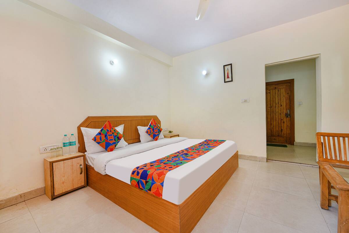 A neatly made bed with a colorful blanket, a wooden nightstand with water bottles, and a wooden bench in a bright room with white walls and a wooden door.