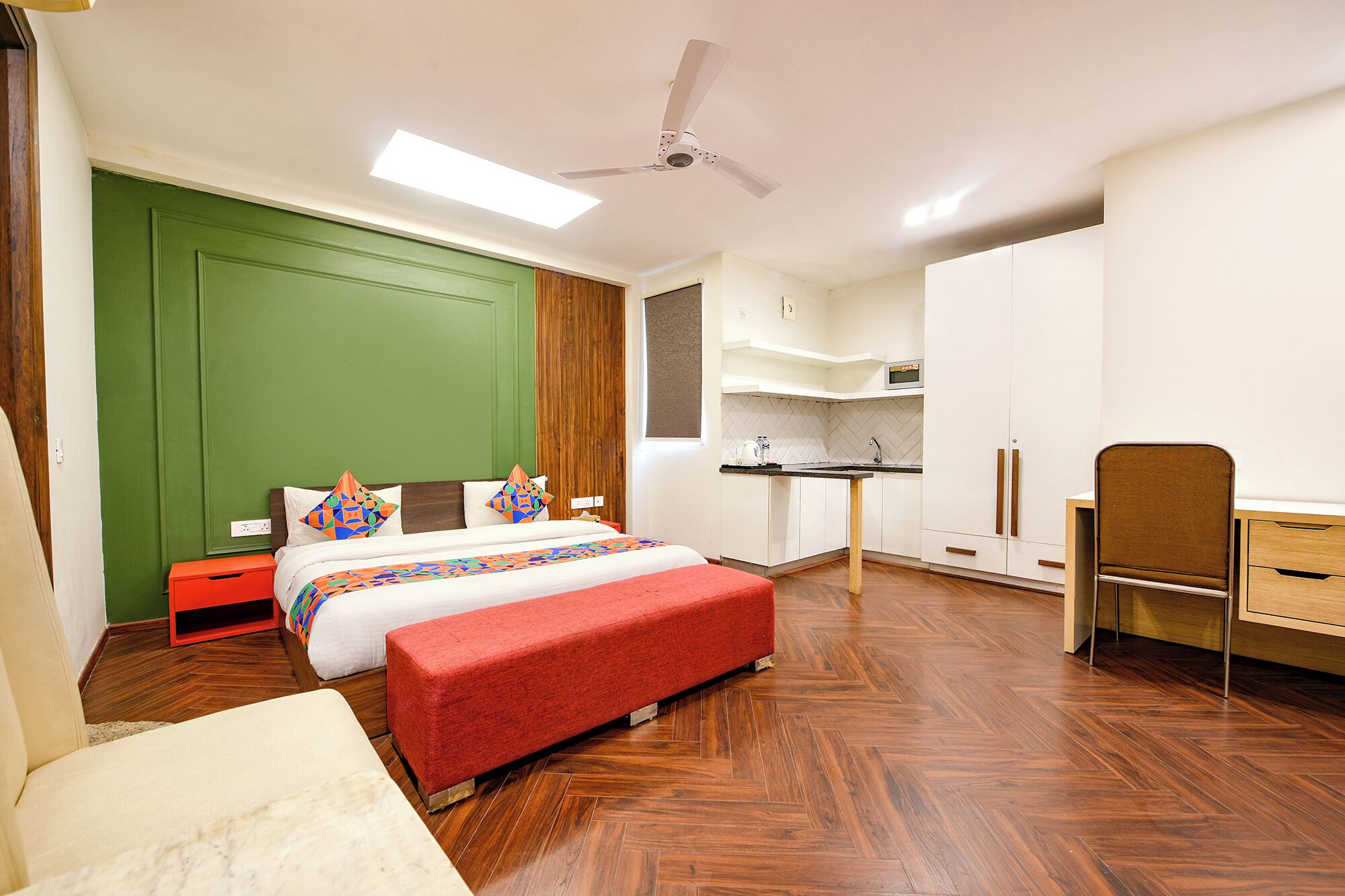 A modern bedroom with a bed, a red bench, a desk, and a kitchenette.