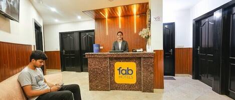 Een hotel receptie met een receptiemedewerker, een gast zittend op een bank en een receptiebalie met het logo van het hotel.