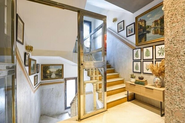 Una escalera con puerta de cristal que da a un nivel inferior, rodeada de obras de arte enmarcadas y una mesa consola de madera.