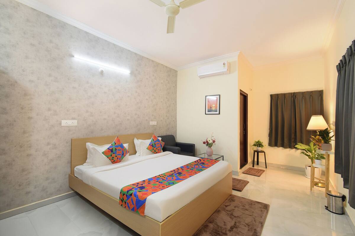 A hotel room with a bed, a bedside table, a lamp, a potted plant, and a view of the outside.