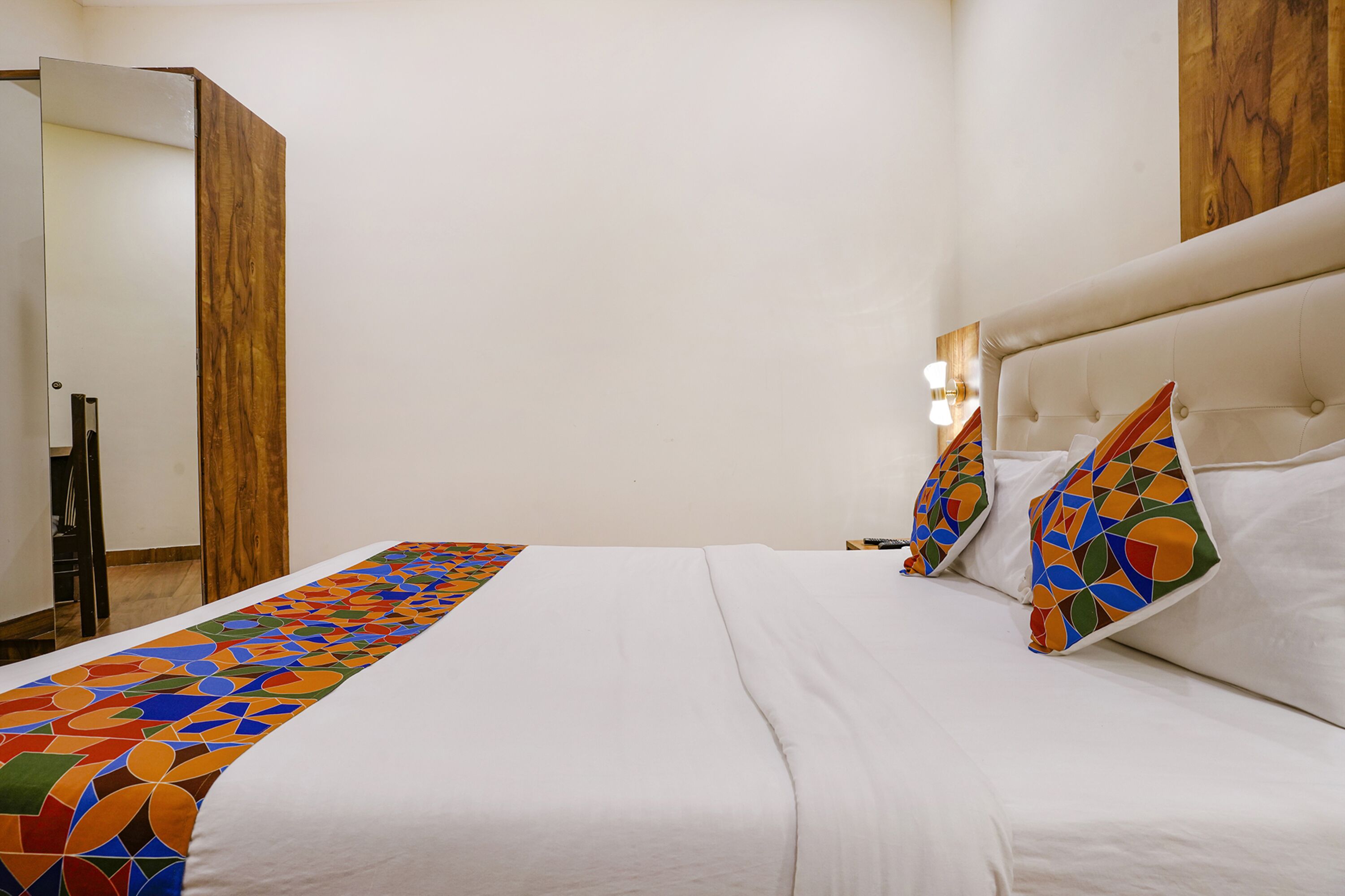 Un lit bien fait, agrémenté d’un couvre-lit aux motifs géométriques colorés, d’une armoire en bois et d’une tête de lit blanche ornée de coussins décoratifs.