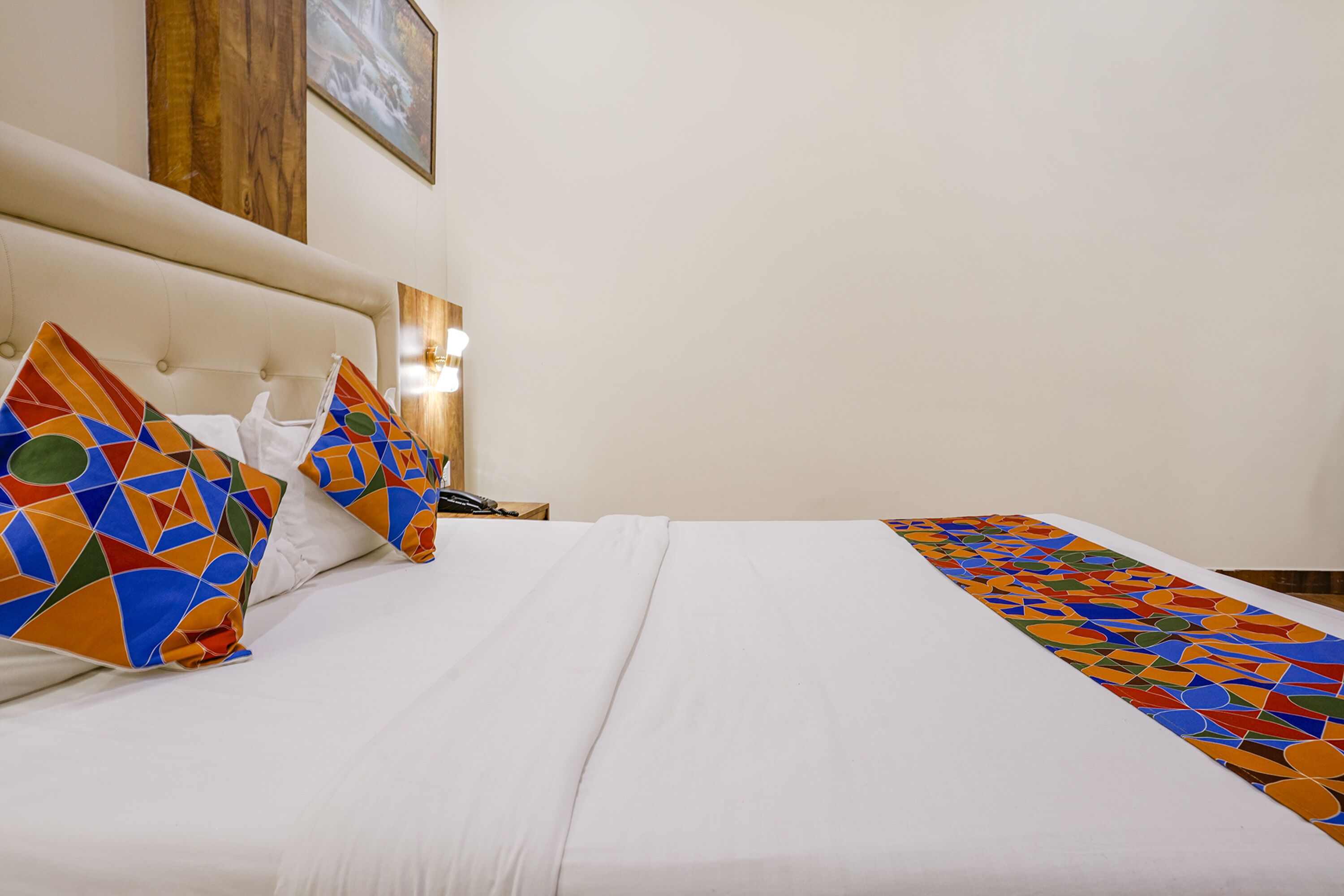 Habitación de hotel con una cama, almohadas de diseño geométrico y colores vivos, una colcha blanca, cabecero de madera y un cuadro enmarcado en la pared.