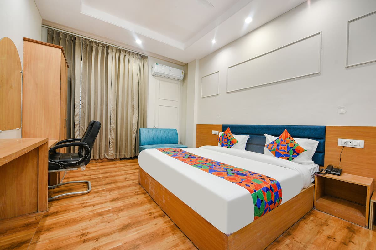 A hotel room with a wooden floor, a bed with a colorful blanket, a desk, a chair, a blue couch, and an air conditioning unit.