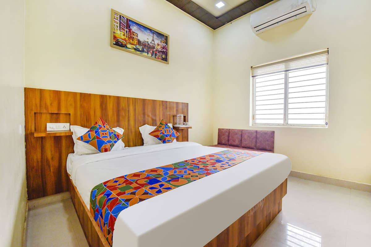 Quarto de hotel com cabeceira de madeira, colcha de cama com padrão geométrico colorido, mesa de cabeceira, janela com persianas e um quadro na parede.