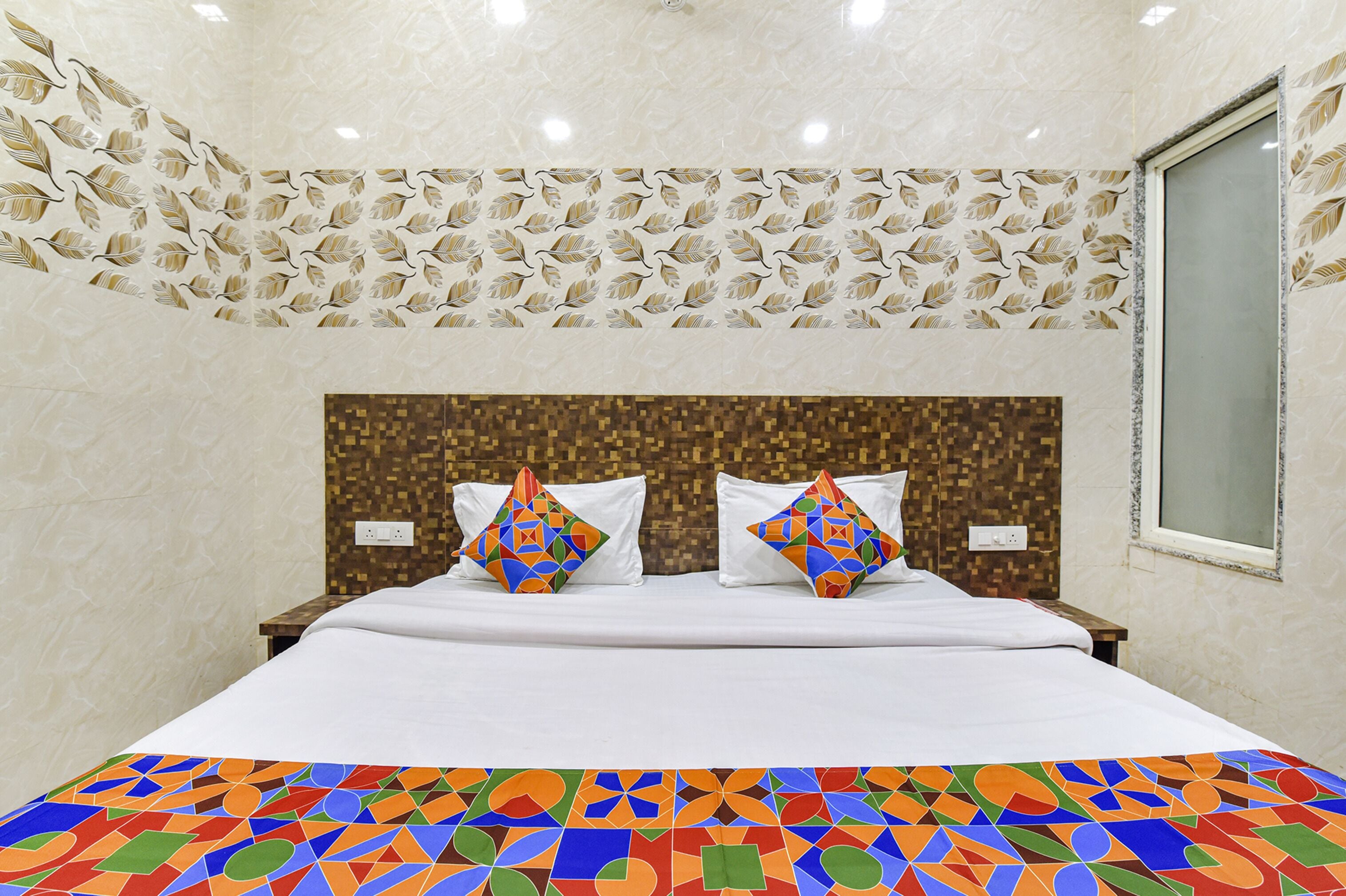 Une chambre d’hôtel avec un grand lit, deux oreillers, une couvre-lit colorée, une tête de lit à motifs et une fenêtre.