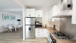 Eine moderne Küche mit weißen Schränken, einem Edelstahlkühlschrank, einer Mikrowelle und einem Gasherd. Im Hintergrund ist ein Essbereich mit Tisch und Stühlen zu sehen.