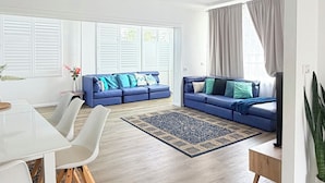 Ein geräumiger Wohnbereich mit weißer Decke, Deckenventilator, blauem Sofagarnitur, Esstisch mit Stühlen und großem Fenster mit Vorhängen.