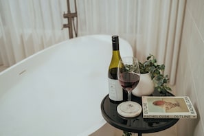 A white bathtub, a bottle of wine, a wine glass, a book titled 'Outline' by Rachel Cusk, and a small potted plant on a black table. - The Homestead: five master suites with ensuites, where luxury meets nature. (Peerabeelup)