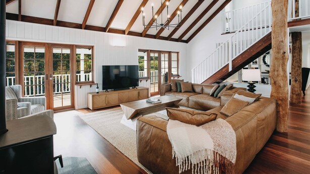 Amplia sala de estar con un sofá modular grande, televisor y chimenea. Se ve una escalera que sube a un nivel superior, y grandes ventanales que dejan entrar luz natural.
