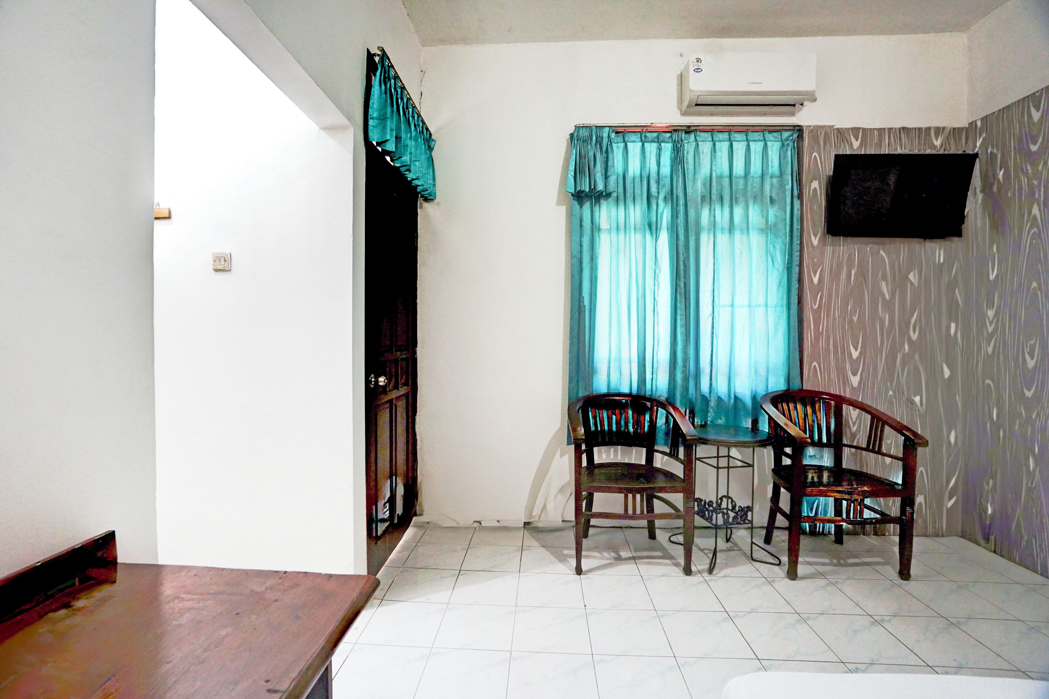 A room with a dining table, two chairs, a TV mounted on the wall, and a window with curtains.