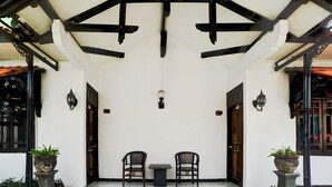 Un espacio al aire libre con techo, amueblado con dos sillas y una mesa, rodeado de paredes blancas y ventanas de marco negro.