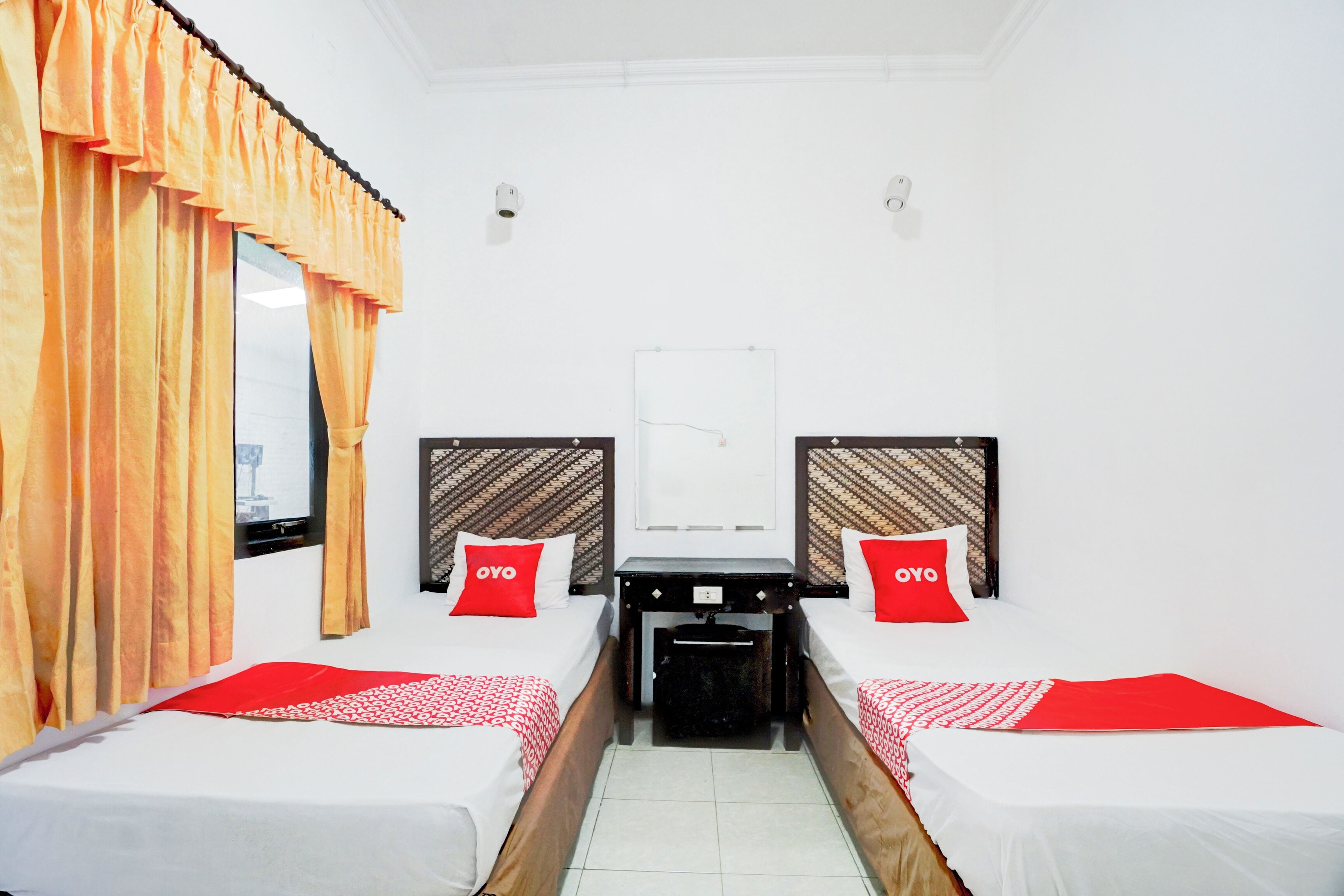 A hotel room with two beds, each with a red pillow, white bedding, and a red and white checkered blanket. There is a small table between the beds, a mirror on the wall, and a window with orange curtains.