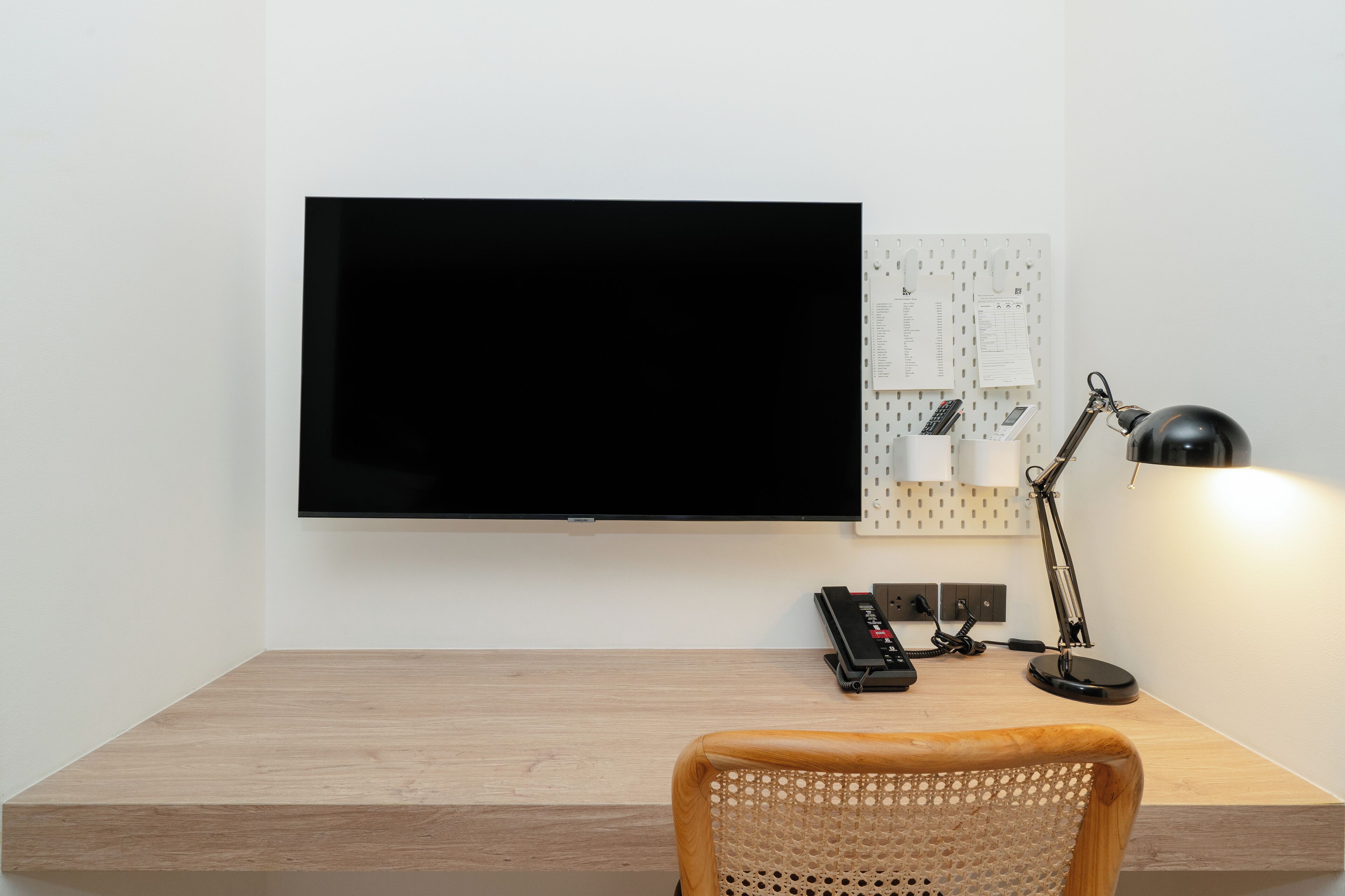 A modern room with a wooden desk, a flat-screen TV mounted on the wall, a desk lamp, a telephone, and a bulletin board with papers.