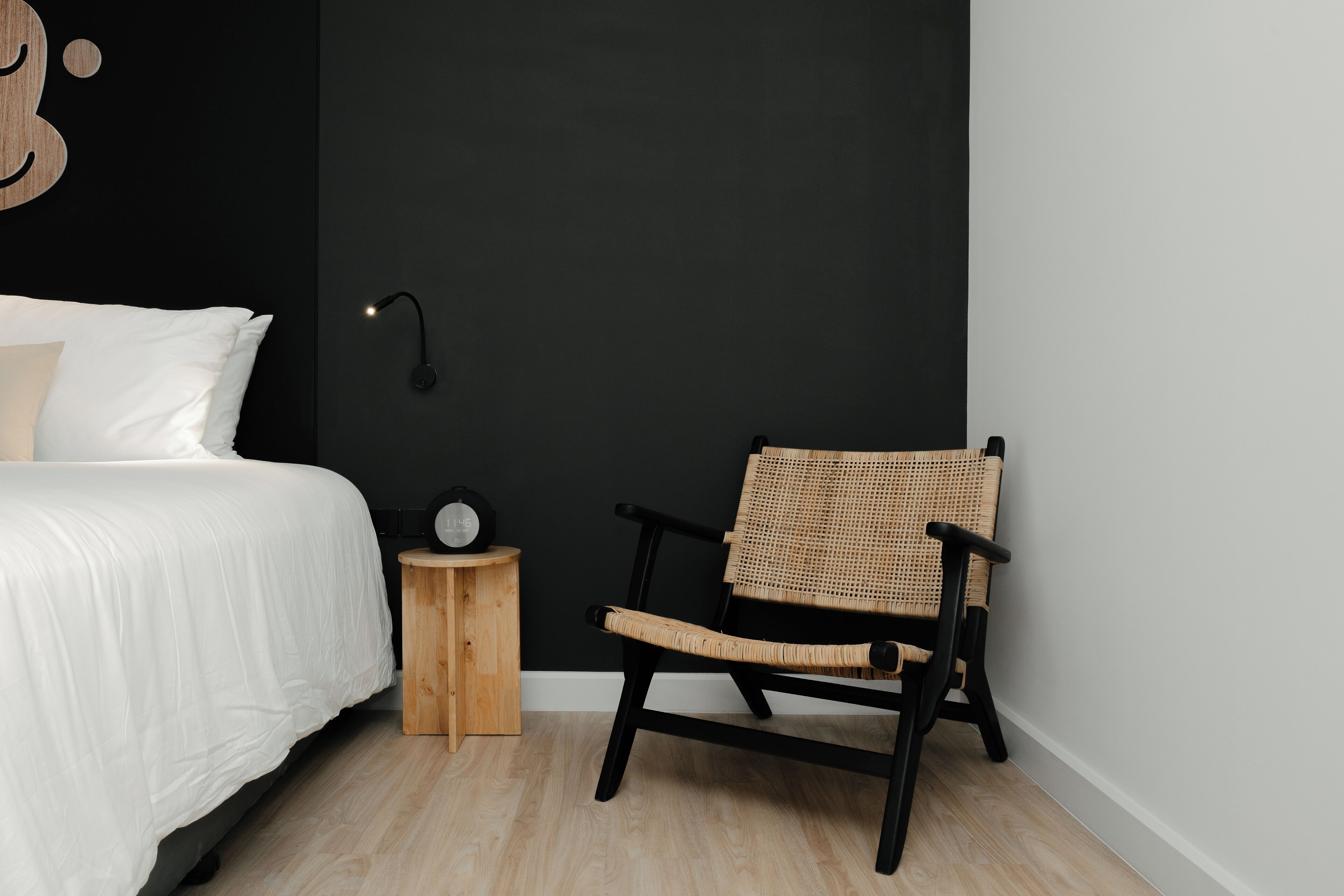 A bedroom with a bed, a wooden nightstand, a black chair, and a wall-mounted lamp.