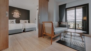 A modern hotel room with a sofa, two beds, a wooden chair, a small table, and a large window offering a city view. - Blu Monkey Hub & Hotel Bangtao Beach Phuket (Choeng Thale)
