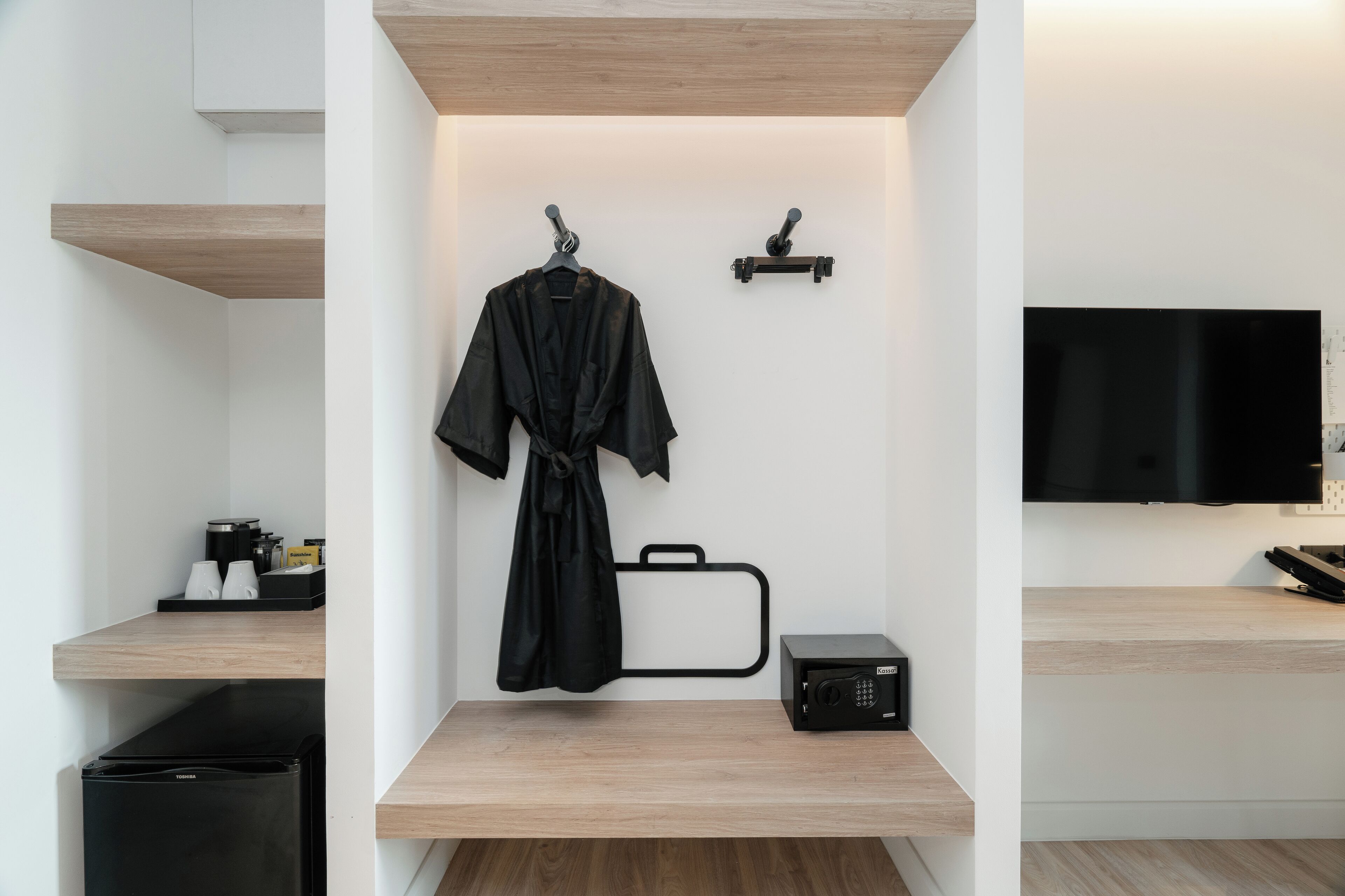 Una camera d'albergo moderna con pavimento in legno, un accappatoio nero appeso a un gancio, una valigia nera su uno scaffale, una TV a schermo piatto montata al muro e un piccolo speaker nero.