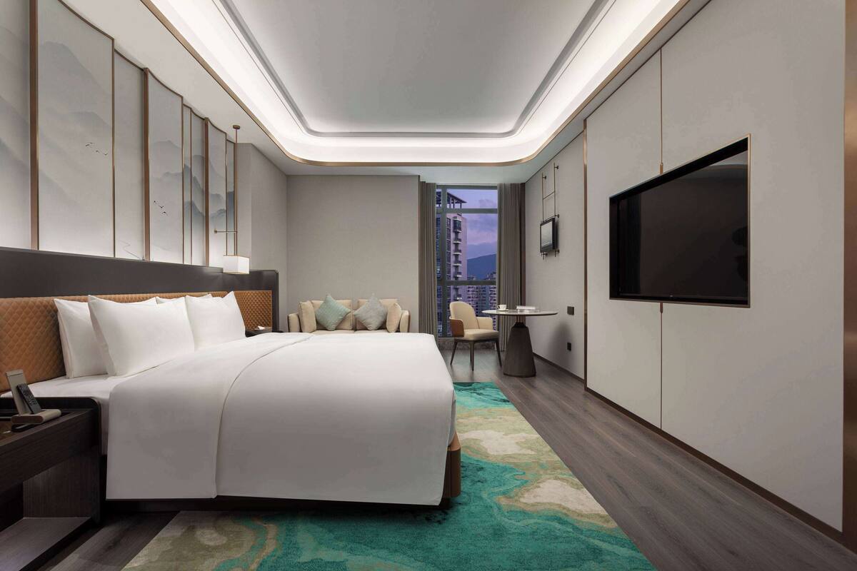Une chambre d’hôtel moderne dotée d’un grand lit, d’un téléviseur à écran plat, d’un bureau avec une chaise et offrant une vue sur le paysage urbain par la fenêtre.