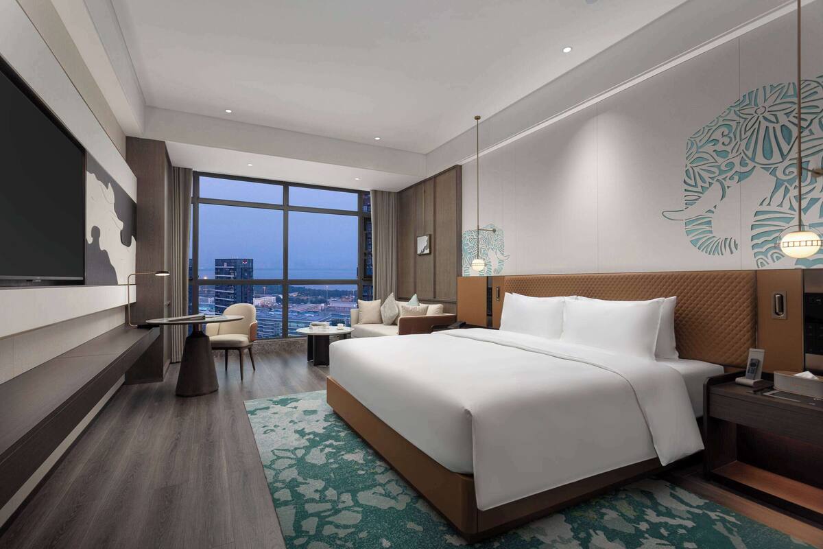 Une chambre d’hôtel moderne dotée d’un grand lit, d’un coin salon avec un canapé et des fauteuils, d’une petite table et d’une grande fenêtre donnant sur la ville.