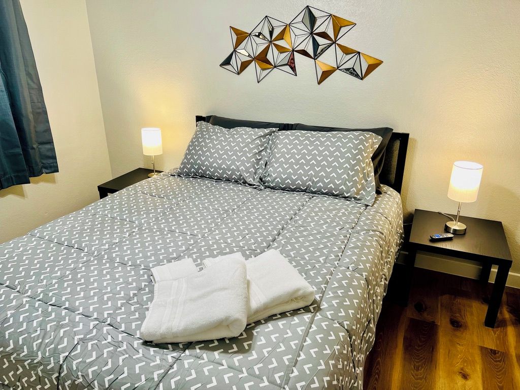 Una cama bien tendida con ropa de cama estampada y una toalla doblada, dos lámparas de noche y un adorno de pared geométrico.