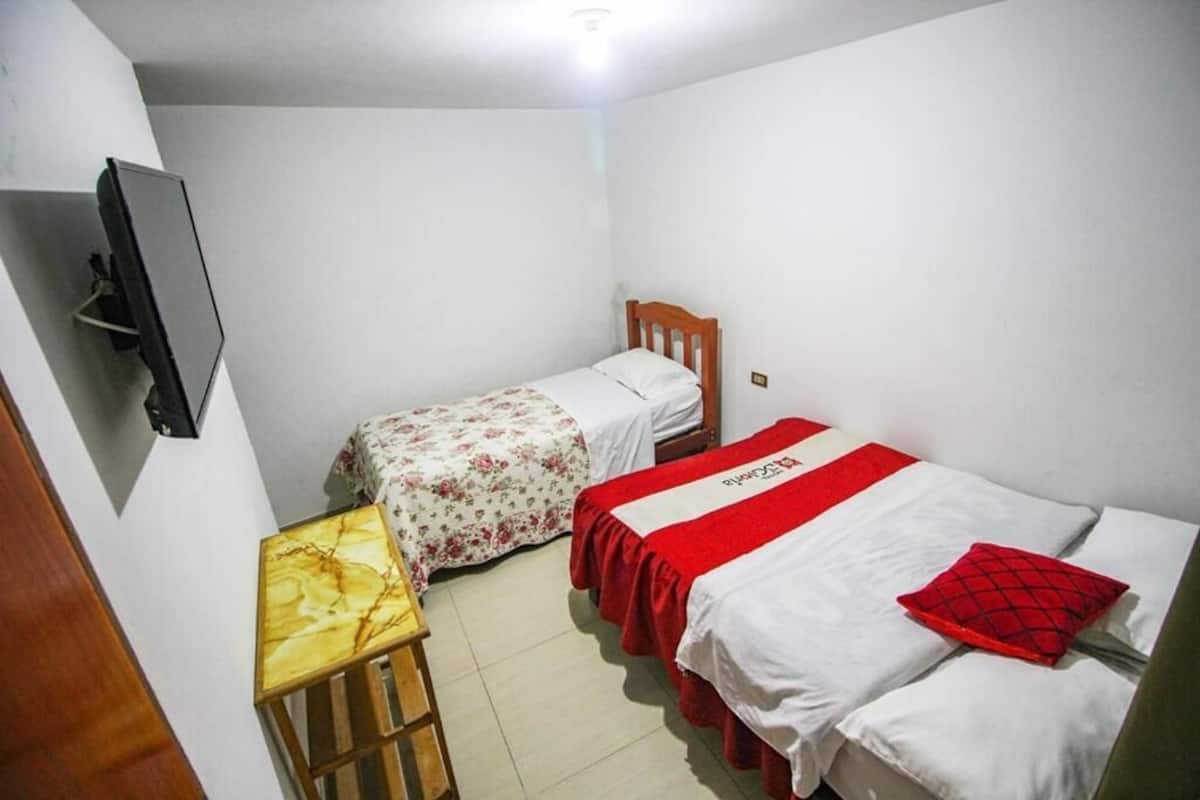 A small, clean room with two single beds, a wooden nightstand, a TV, and a red pillow.