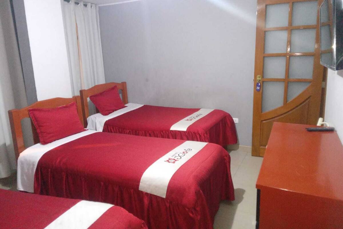 A hotel room with two beds, each with red bedding and white accents, a wooden door, and a small desk with a remote control.