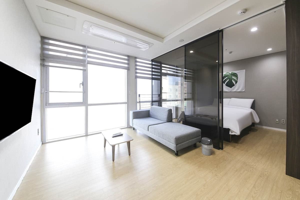 Een moderne hotelkamer met een grijze bank, een tafeltje, een bed met witte lakens en een groot raam met zonwering.