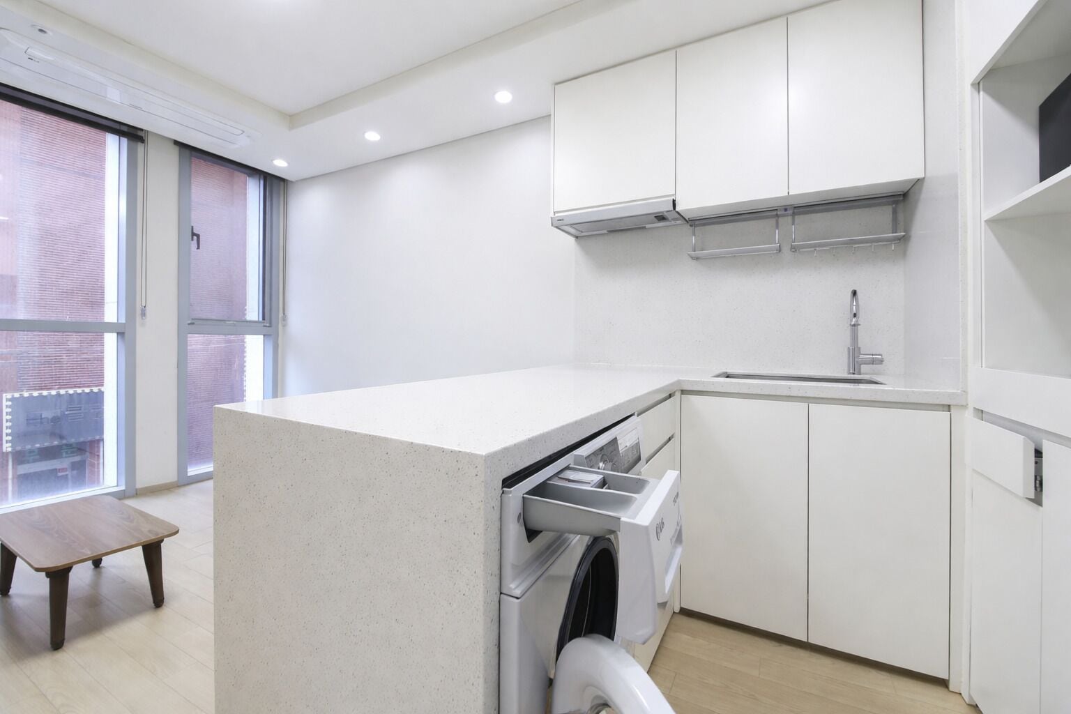 A modern kitchen with a built-in washer and dryer, white cabinetry, and a large window providing natural light.