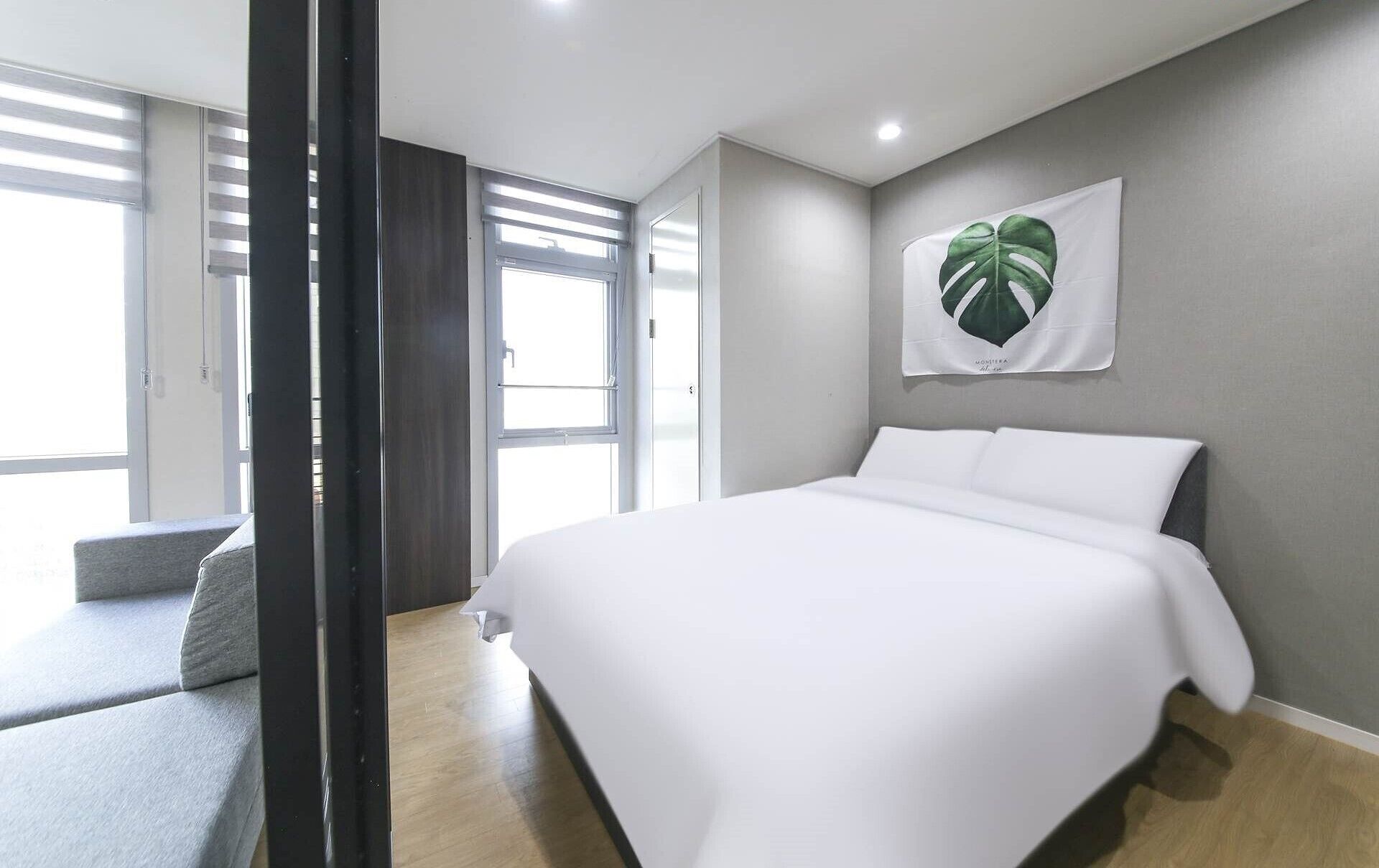 Une chambre moderne avec un grand lit, une fenêtre avec des stores, une plante fixée au mur et un canapé gris.