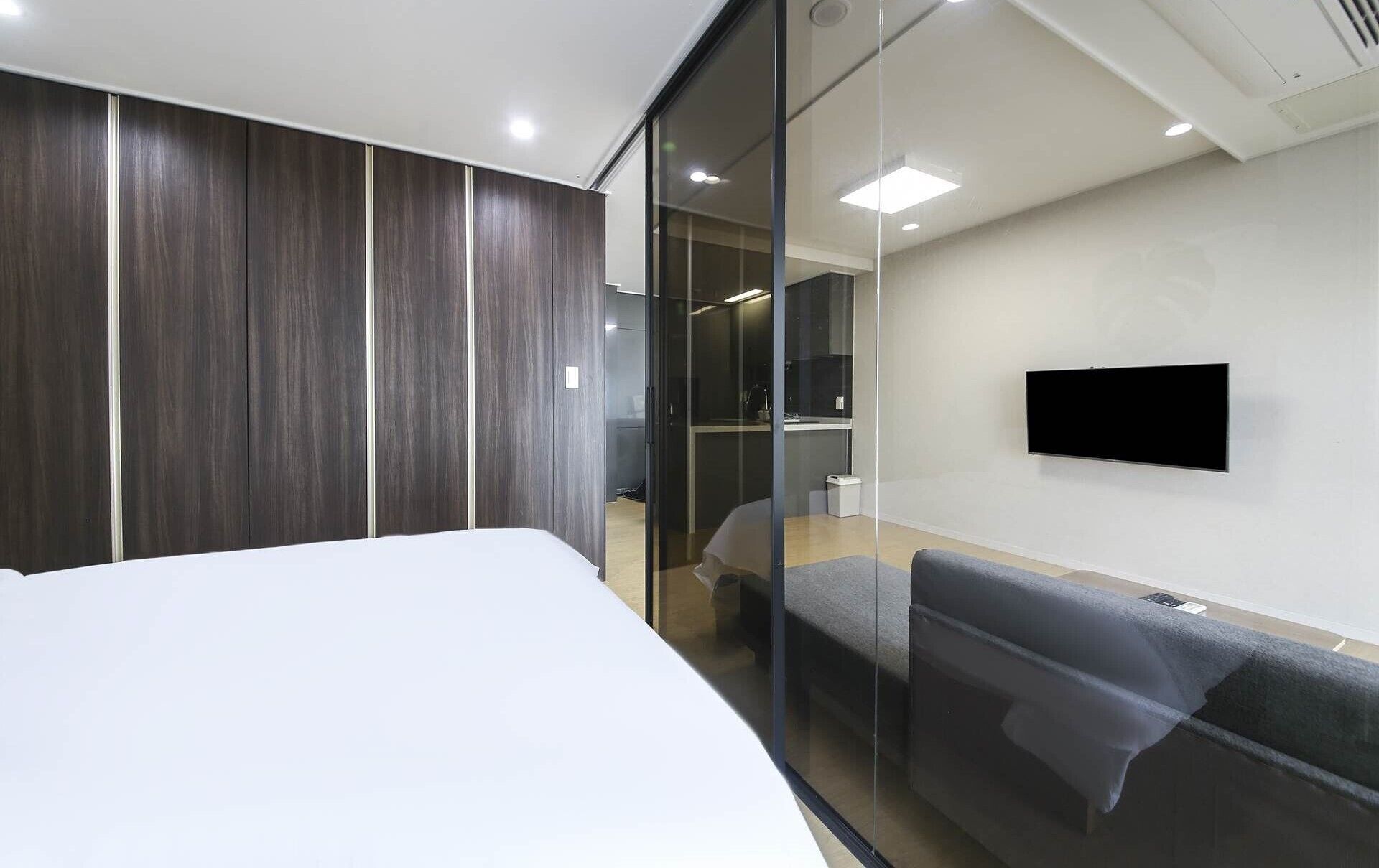 A modern hotel room with a large bed, a sofa, a flat-screen TV mounted on the wall, and a glass partition separating the room from the hallway.