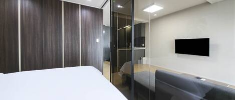 A modern hotel room with a large bed, a sofa, a flat-screen TV mounted on the wall, and a glass partition separating the room from the hallway.