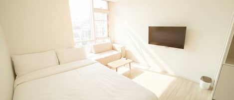 Una habitación de hotel moderna con una cama grande, un televisor de pantalla plana montado en la pared, una pequeña zona de estar con un sofá y una mesa, y un ventanal que proporciona luz natural.
