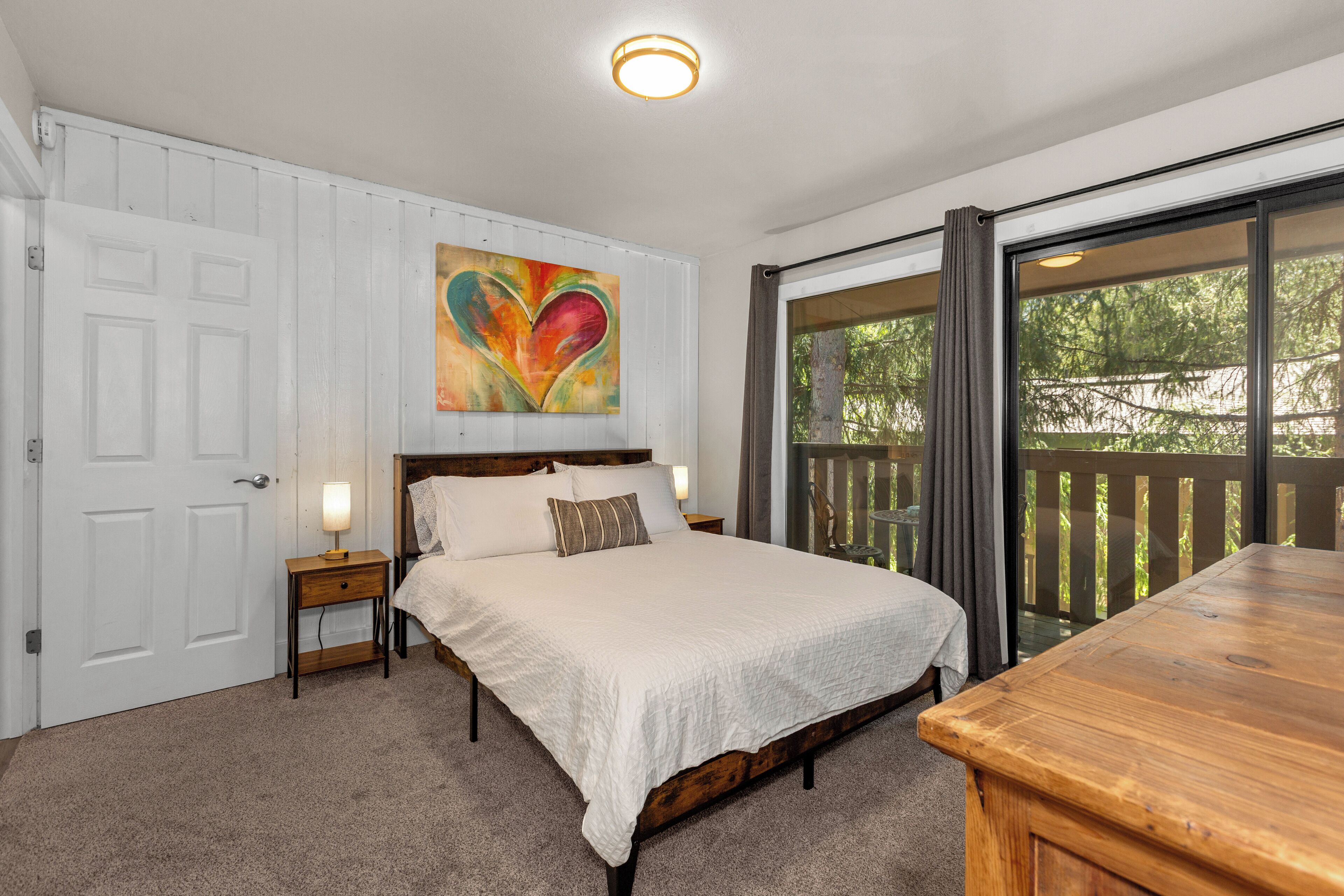 A bedroom with a large bed, a nightstand, a painting above the bed, a window with a view of trees, and a wooden table.