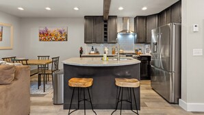 A modern kitchen with dark cabinets, a stainless steel refrigerator, and a central island with stools. - Stylish Sun Valley 2BR - Dog-Friendly - 2 Min Walk to Town (Ketchum)