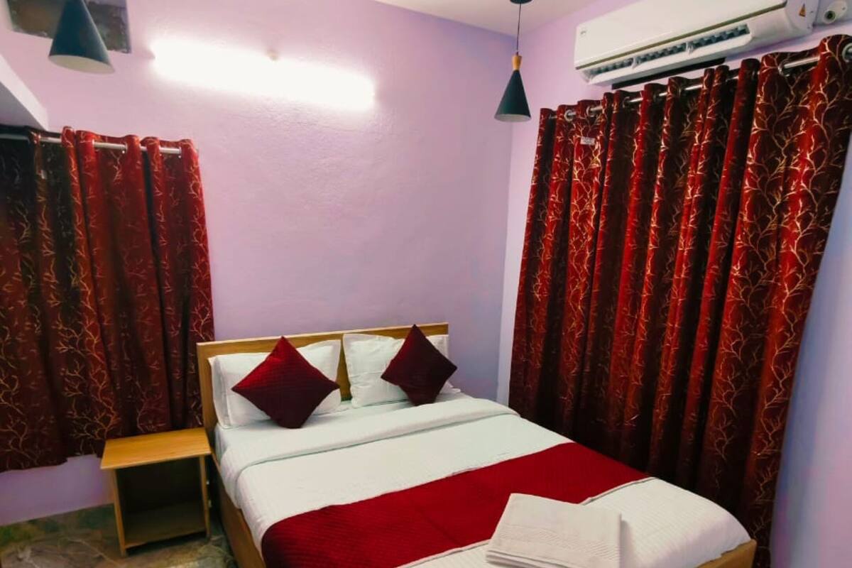 A neatly made bed with a red and white bedspread, two pillows, and a folded towel on the side. There is a wooden nightstand beside the bed, and a small table with a lamp. The room has red curtains with a leaf pattern, and an air conditioning unit is mounted on the wall.