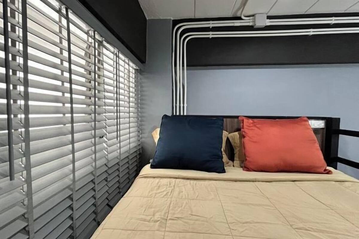 A bedroom with a bed, two pillows, a window with blinds, and visible plumbing on the wall.