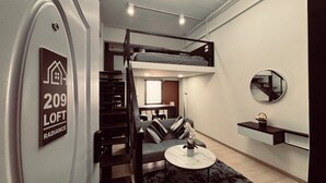 A loft-style room with a bunk bed, a small seating area, a round mirror, and a sign indicating the room number and type. - The Loft Living Space (Samut Prakan)
