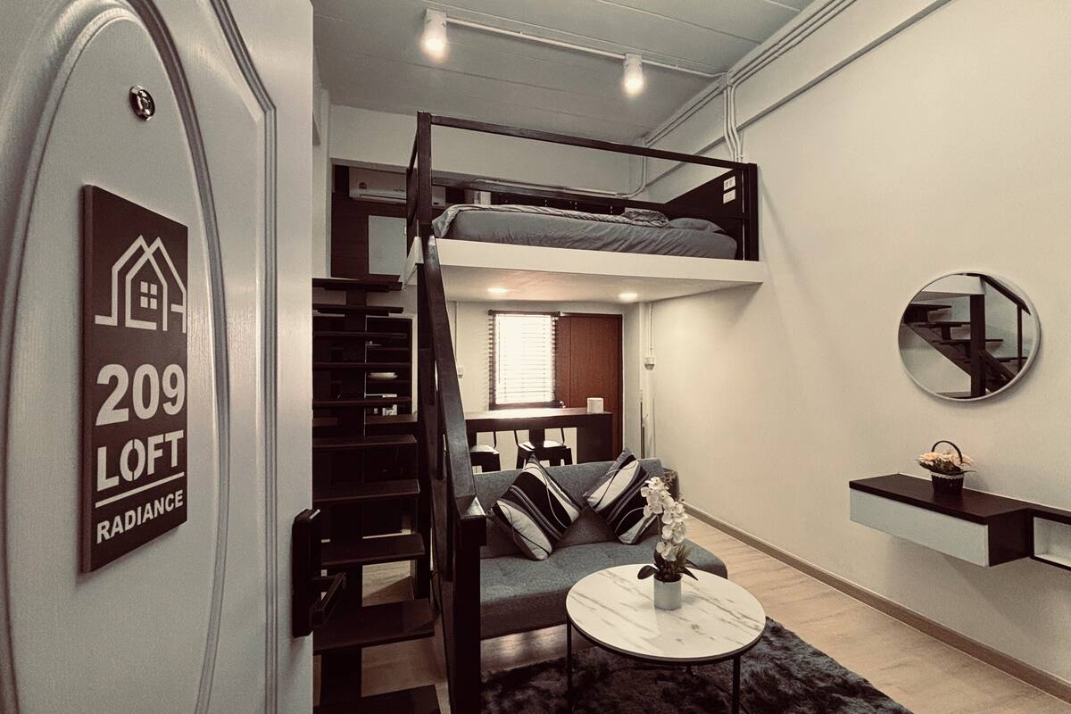 A loft-style room with a bunk bed, a small seating area, a round mirror, and a sign indicating the room number and type.