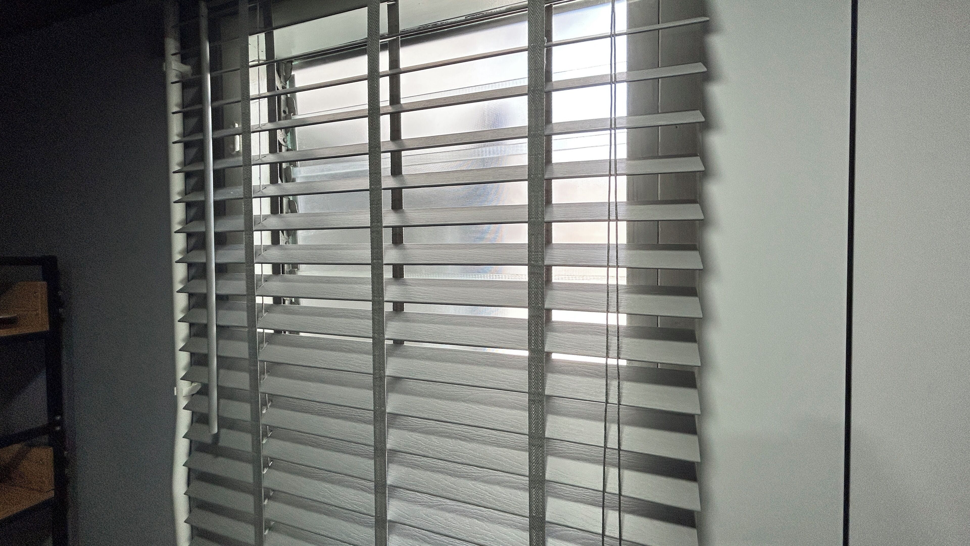 A room with a window covered by horizontal blinds, a wooden shelf, and a partial view of a wall.