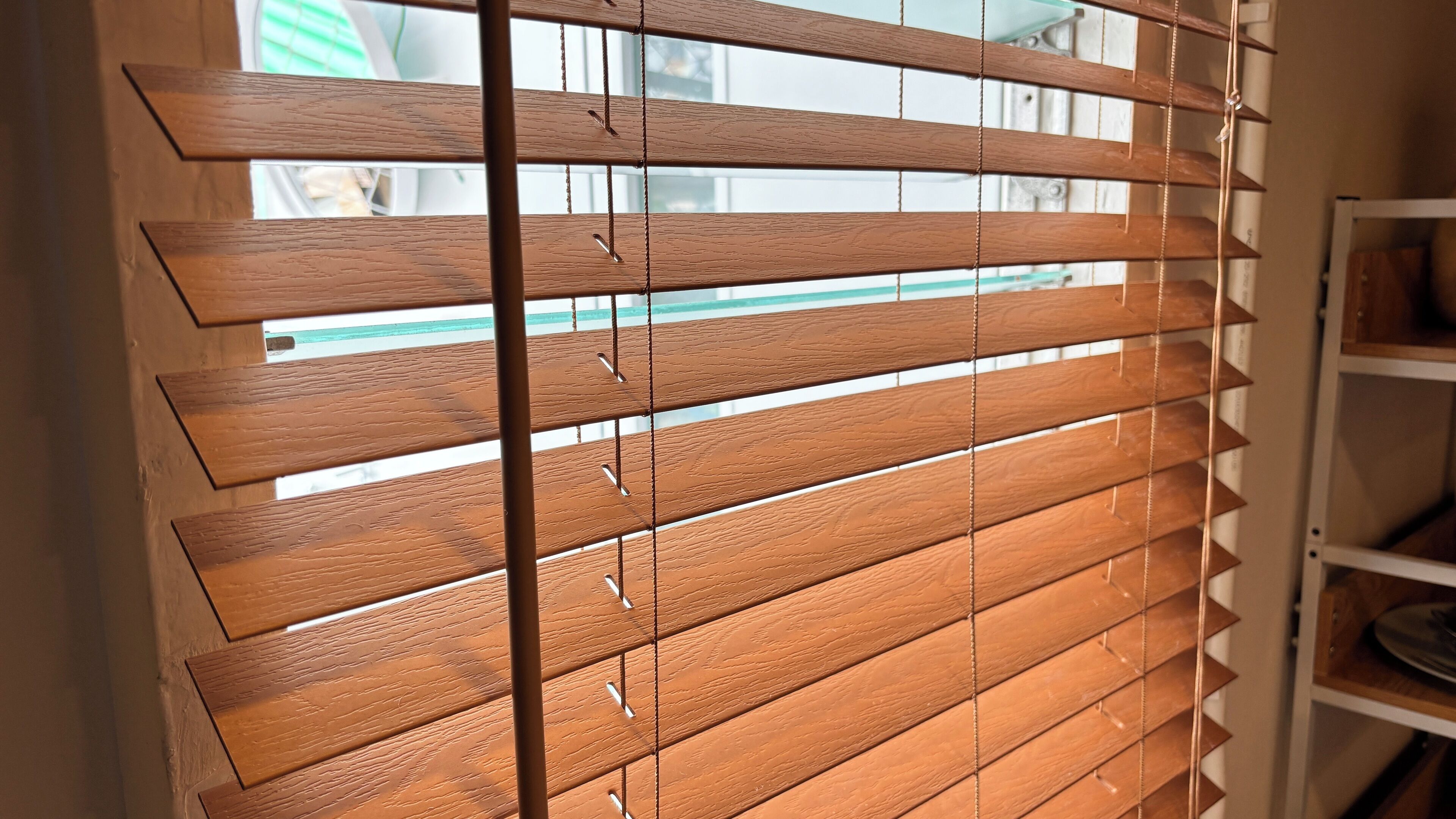 A room with wooden blinds partially open, revealing a view of a window and a shelf with books.