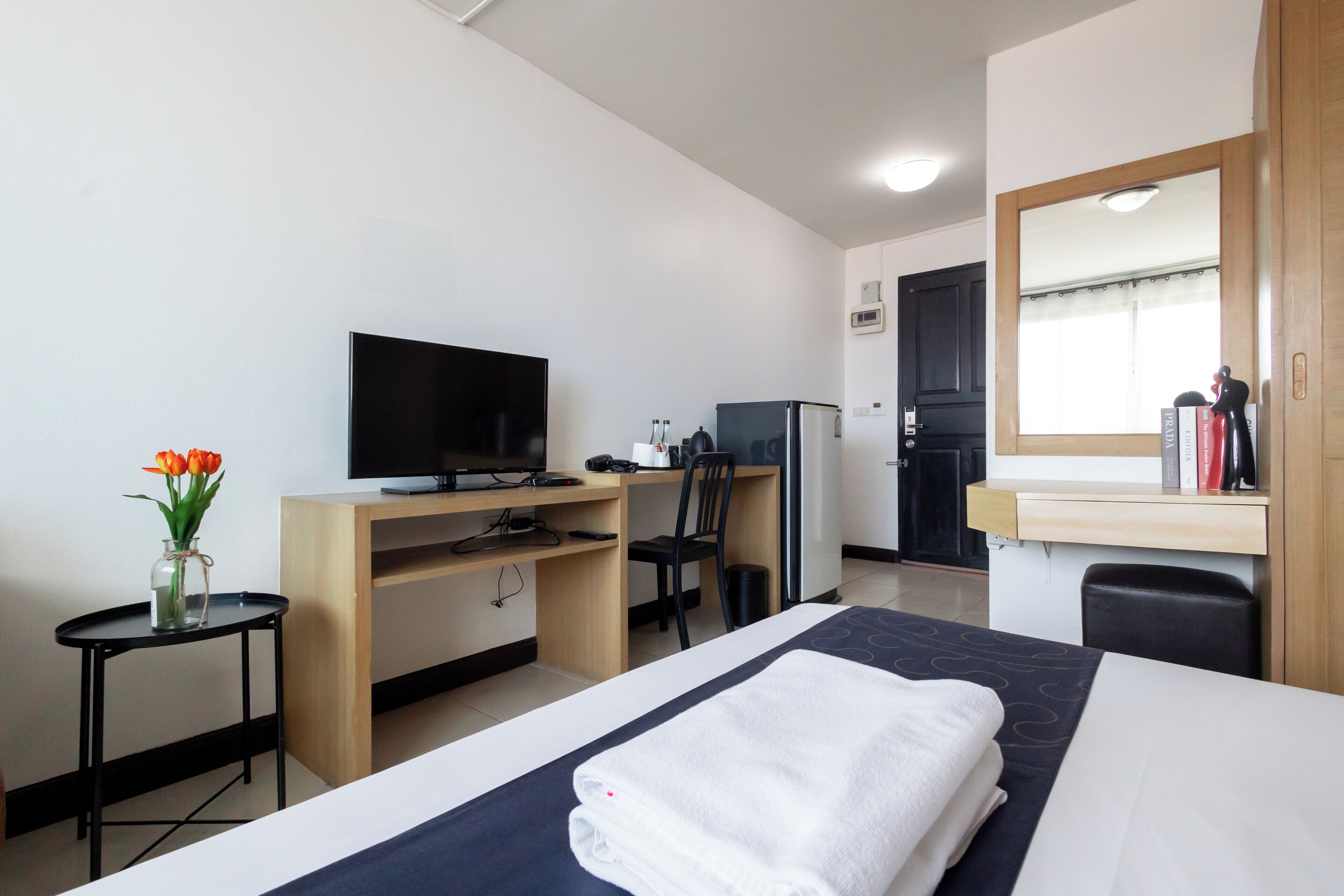 A hotel room with a bed, a desk with a TV, a chair, a small table with a vase of flowers, and a mirror.