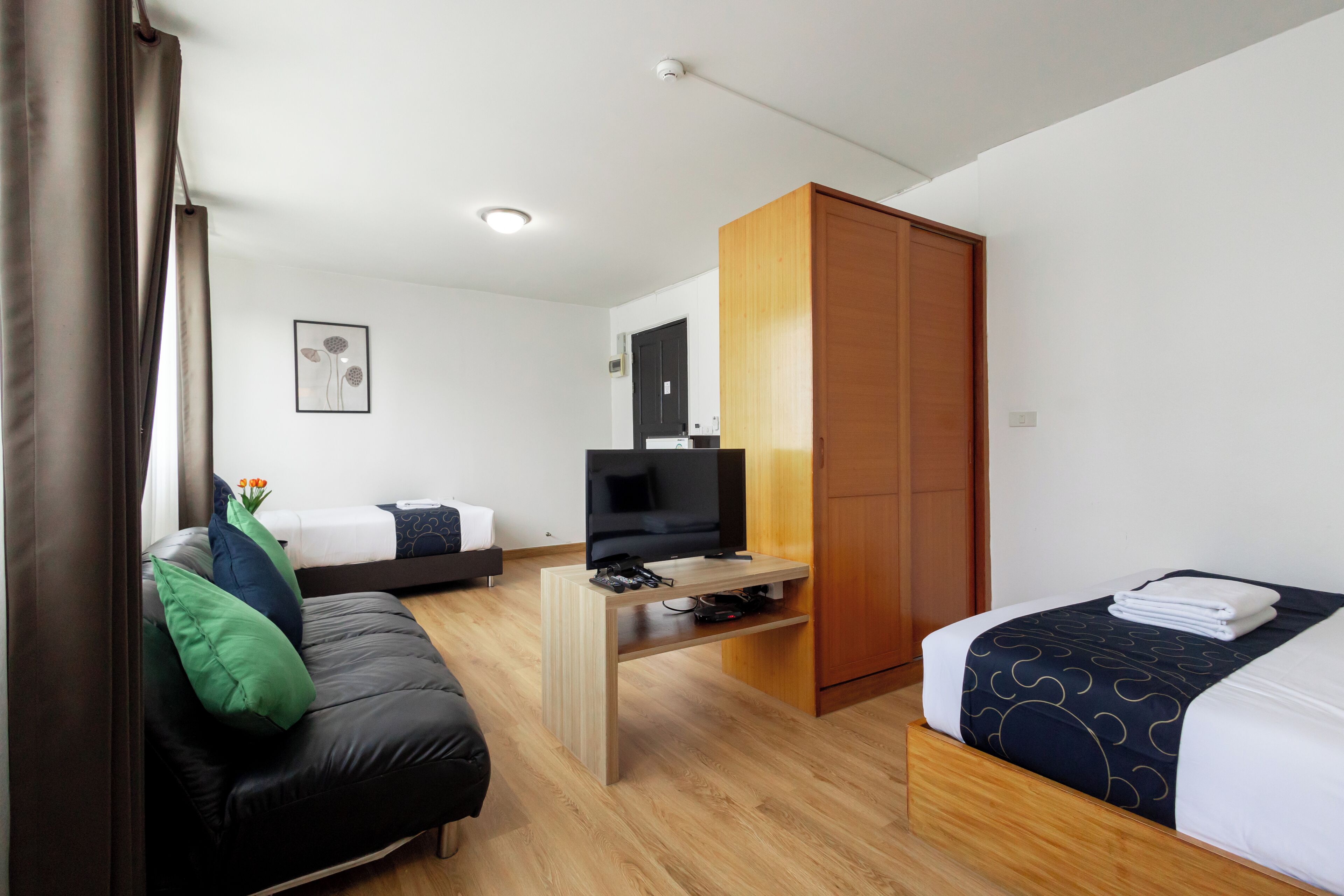 A modern hotel room with a wooden floor, a black sofa with green and blue cushions, a bed with a dark patterned bedspread, a wooden wardrobe, and a television on a wooden stand.